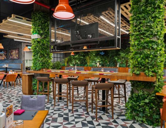 Modern restaurant interior with wooden tables, chairs, and a bar counter, surrounded by green plants and decorative lighting.