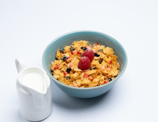 Blue bowl filled with cornflakes, topped with raspberries and blueberries, with a small pitcher of milk on the side.