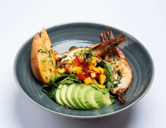 A plate of grilled prawns served with a mango salsa, avocado slices, and garlic bread.