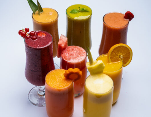 A collection of colorful smoothies and juices in tall glasses, topped with fruits and herbs.