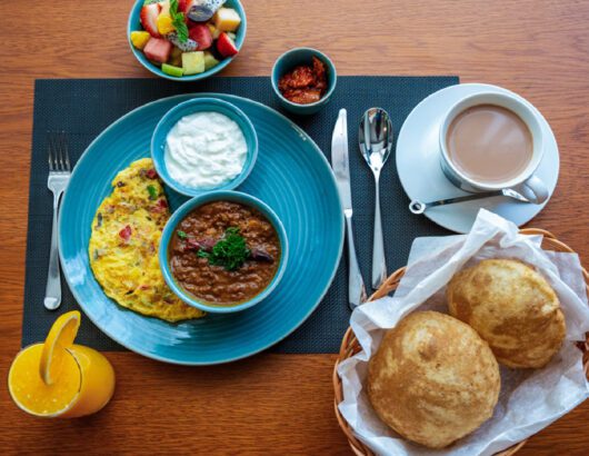A breakfast platter with a vegetable omelette, lentil curry, fruit salad, yogurt, bread, and a cup of tea.