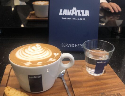 A latte art coffee with a heart design, served on a wooden tray with a glass of water and a small plate of biscuits.