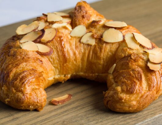 A freshly baked almond croissant with sliced almonds on top, resting on a wooden cutting board.